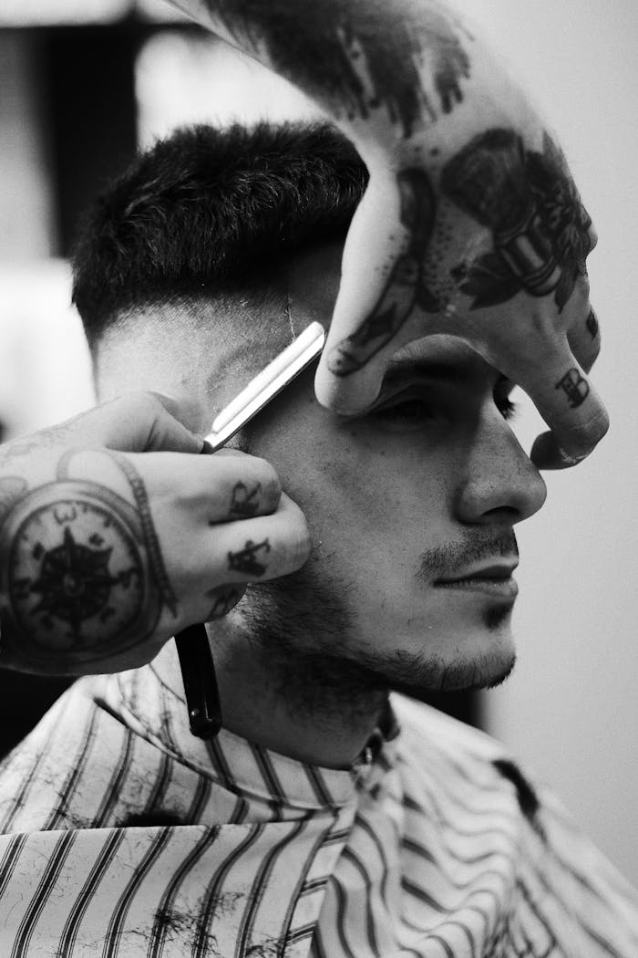 Black and white image showcasing a tattooed barber skillfully shaving a man's sideburns indoors.